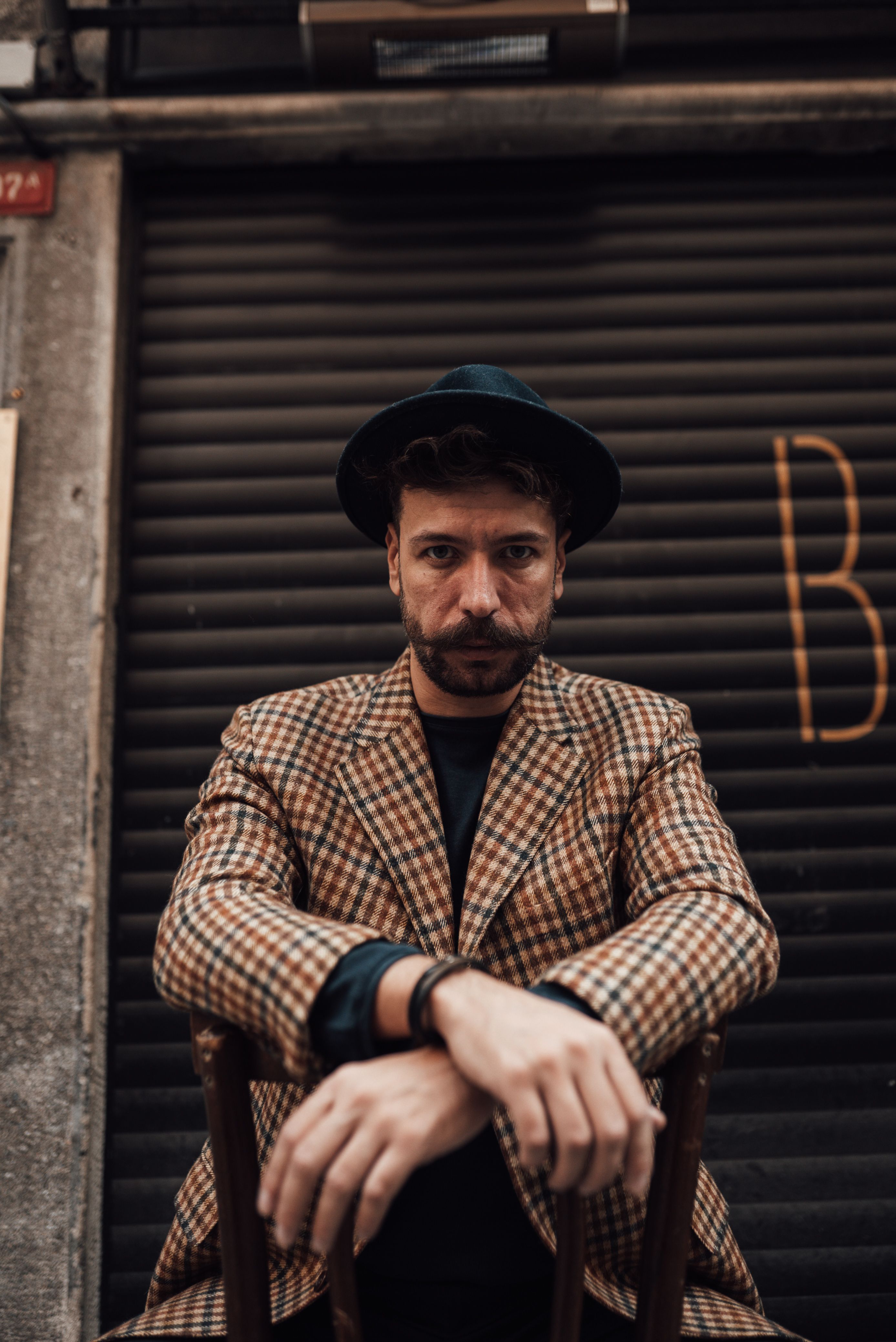 Low angle of bearded male in trendy jacket and hat sitting on chair with crossed arms while looking at camera