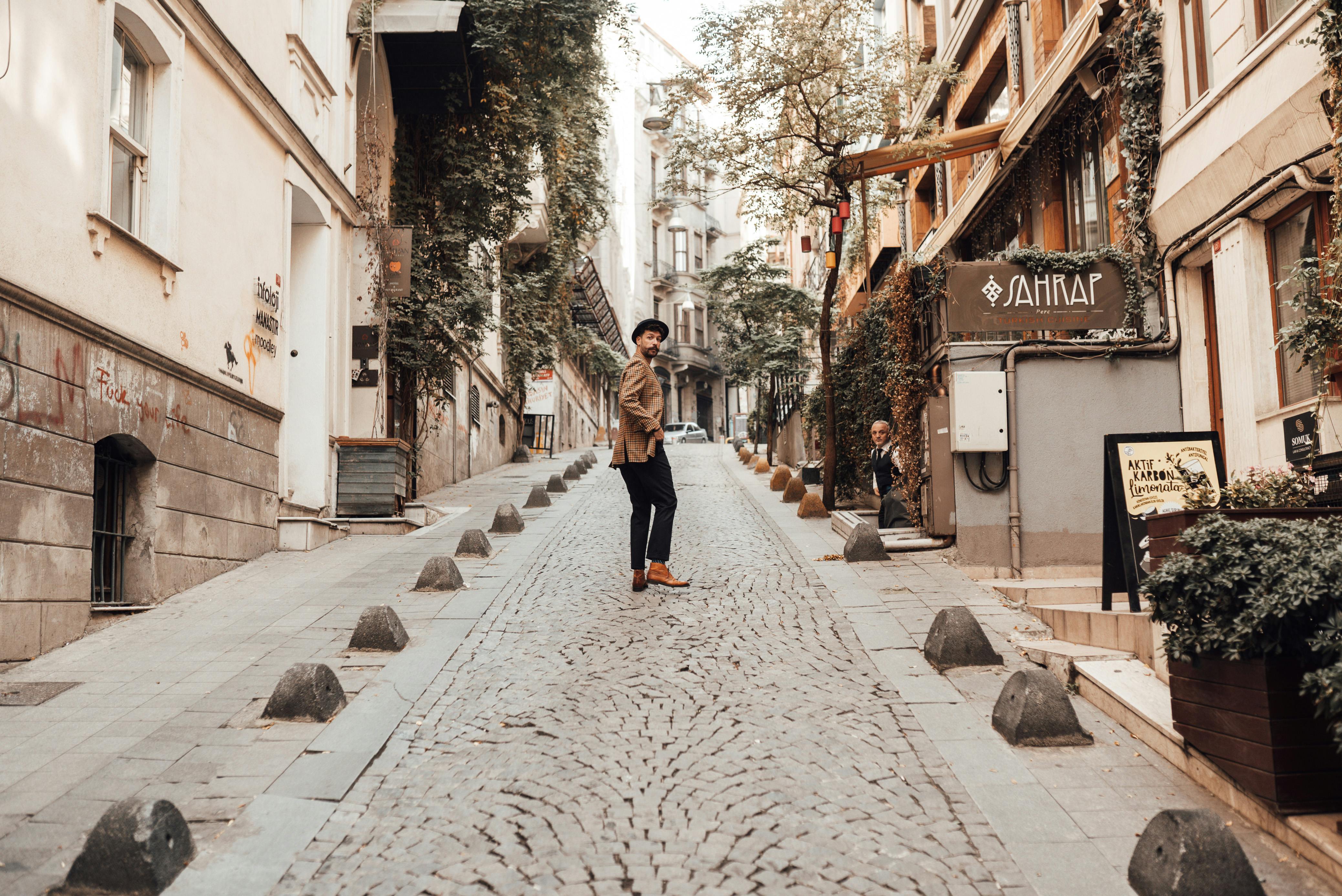 Full body of bearded male in trendy outfit standing on cobblestone walkway between buildings with hand in pocket and looking over shoulder