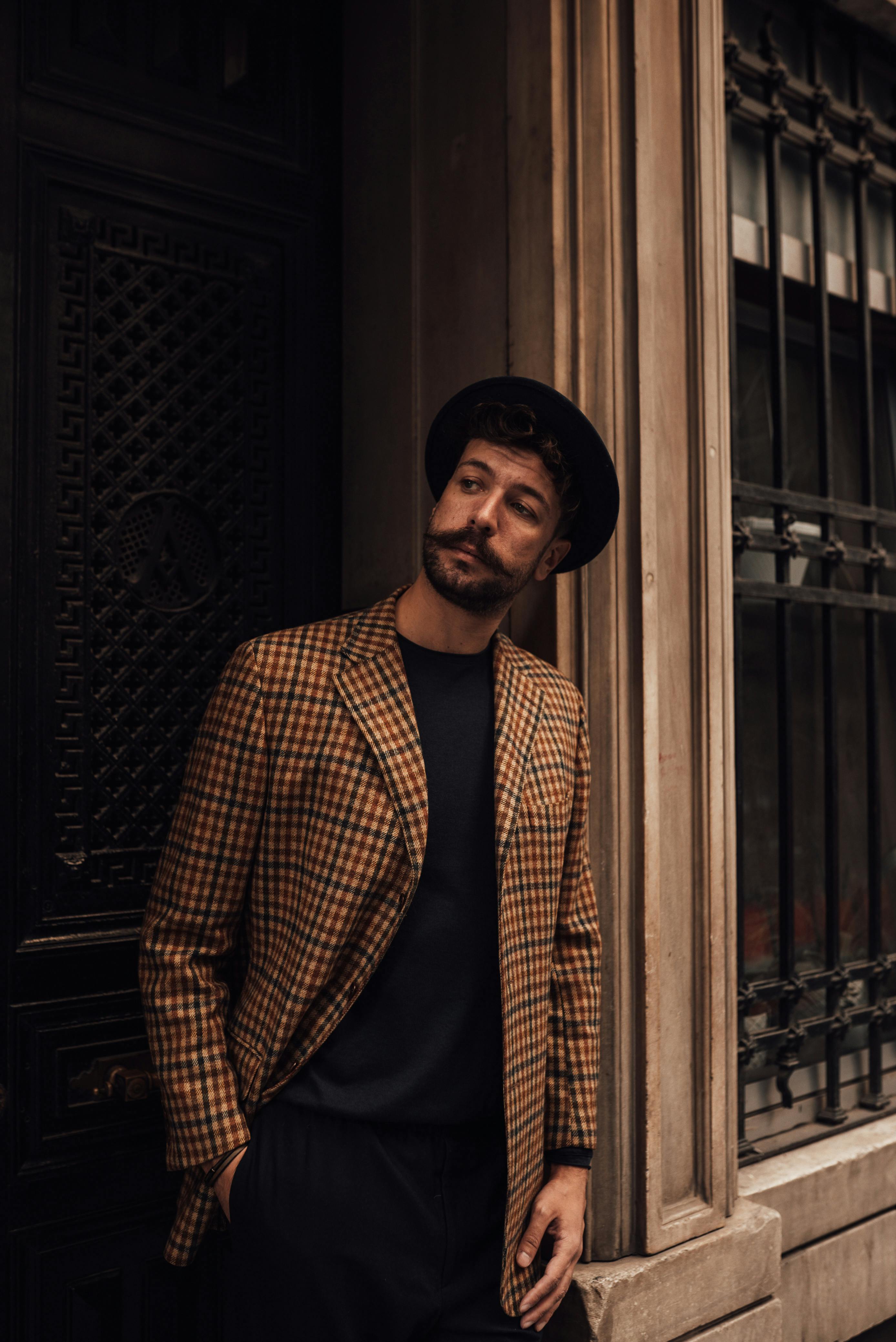 Confident bearded male in trendy hat and jacket leaning on wall with hand in pocket while thoughtfully looking away