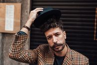 Handsome male model with trendy haircut and beard taking off hat while standing near building and looking at camera