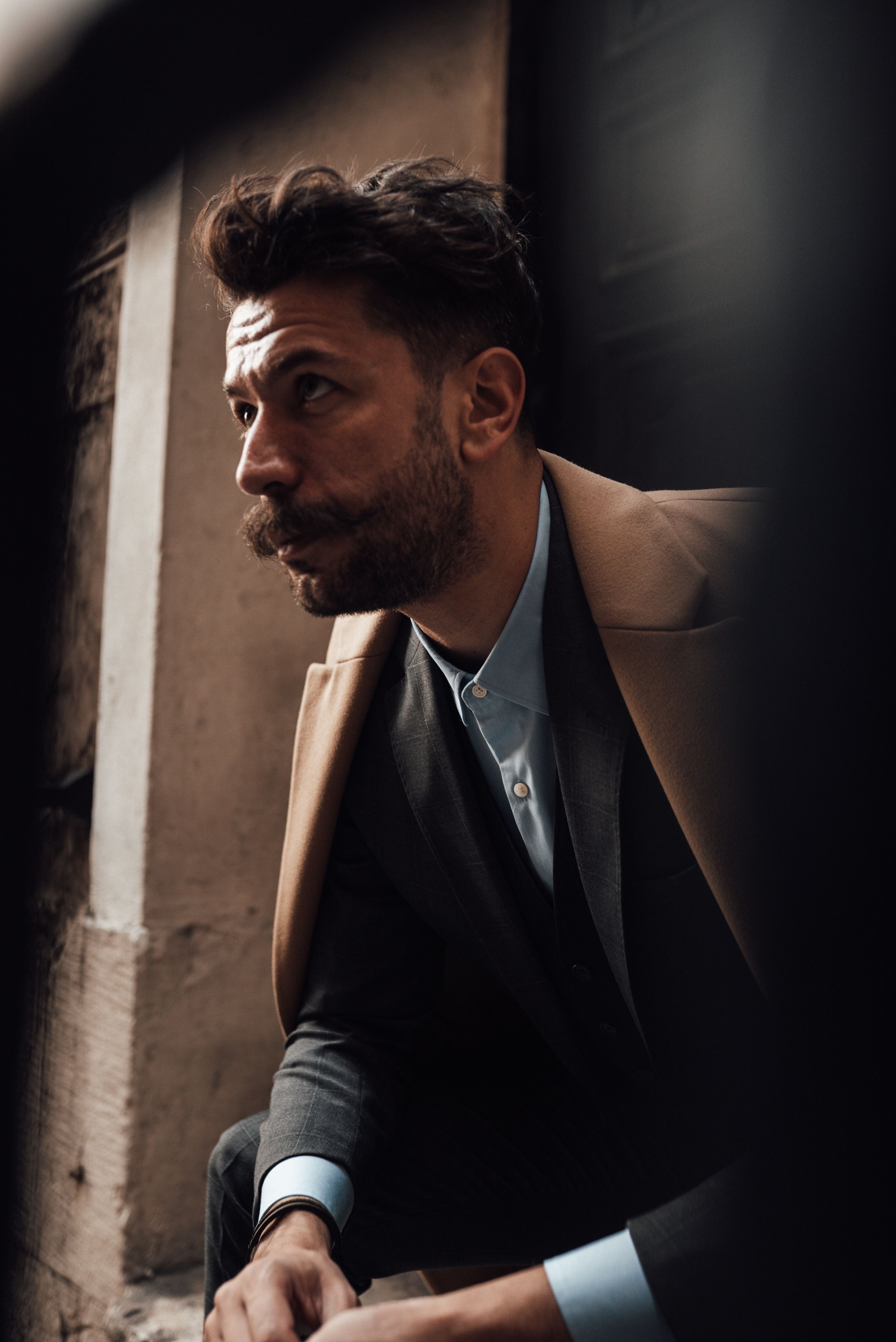 Pensive man with dark hair and mustache in formal clothes sitting on stairs of building and thoughtfully looking away in daytime