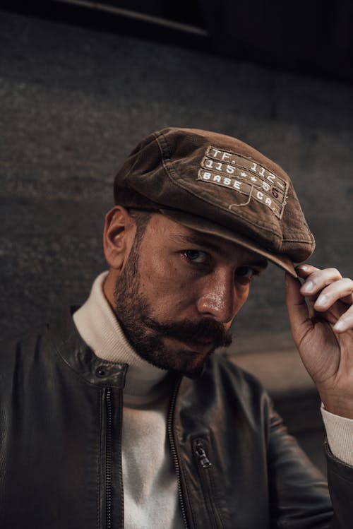 Confident bearded male model with mustache in black leather jacket touching hat while looking at camera