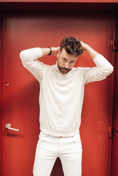 Handsome bearded male model in total white clothes standing near red door and looking at camera
