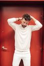 Handsome bearded male model in total white clothes standing near red door and looking at camera