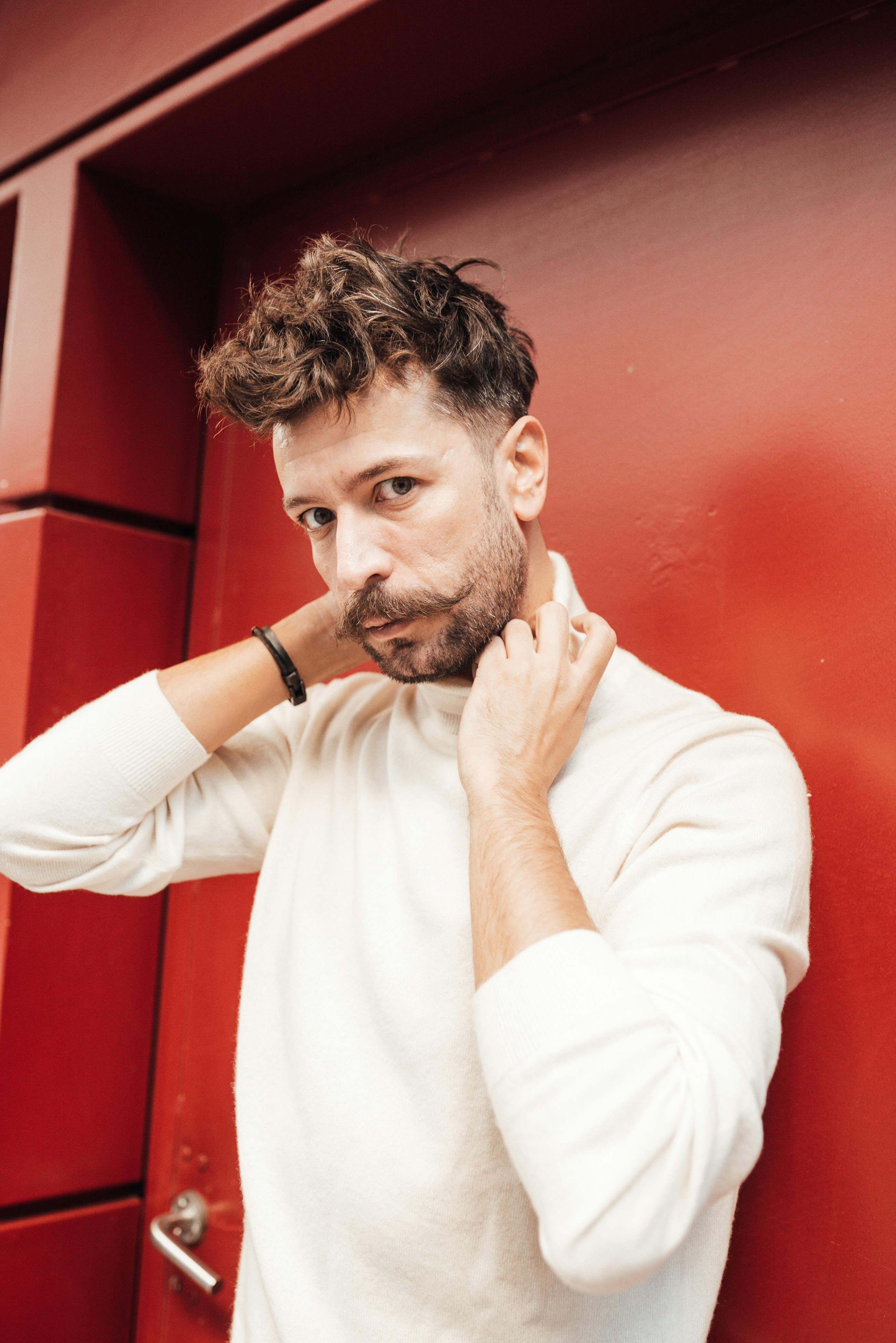 Confident bearded male model in knitted turtleneck with trendy hairstyle holding hands on neck while looking at camera