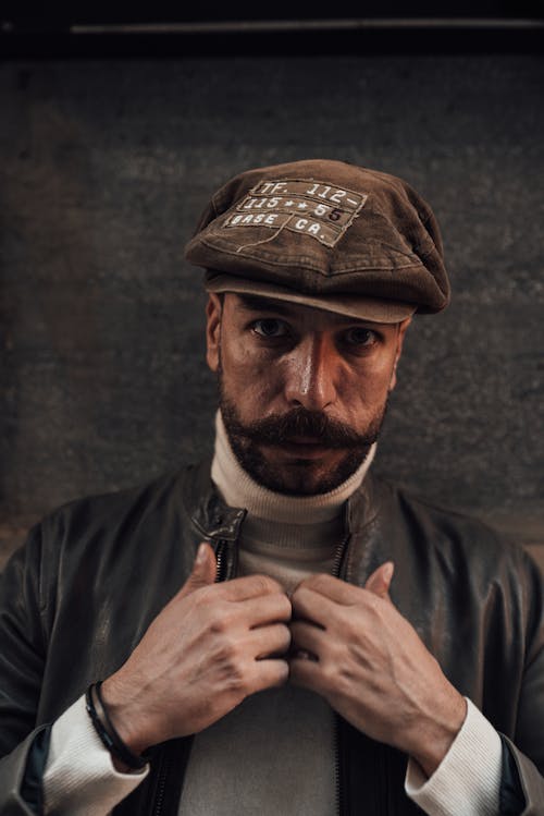 Confident bearded male with mustache in black leather jacket and hat looking at camera