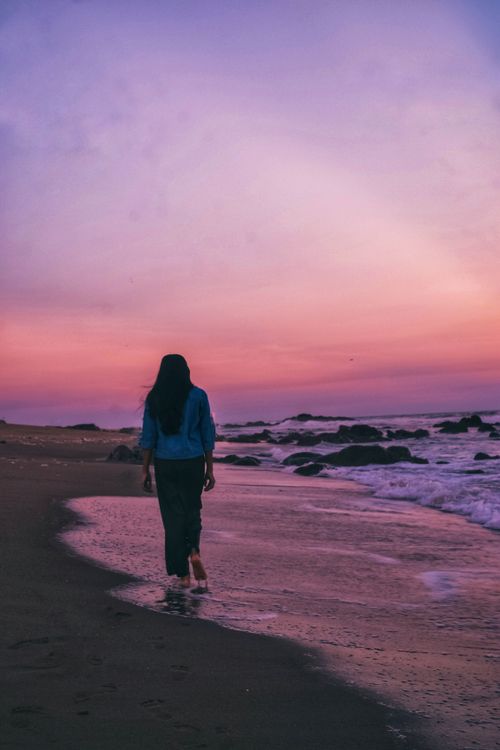Free A woman walks along a sandy beach during a colorful sunset, creating a peaceful and tranquil scene. Stock Photo