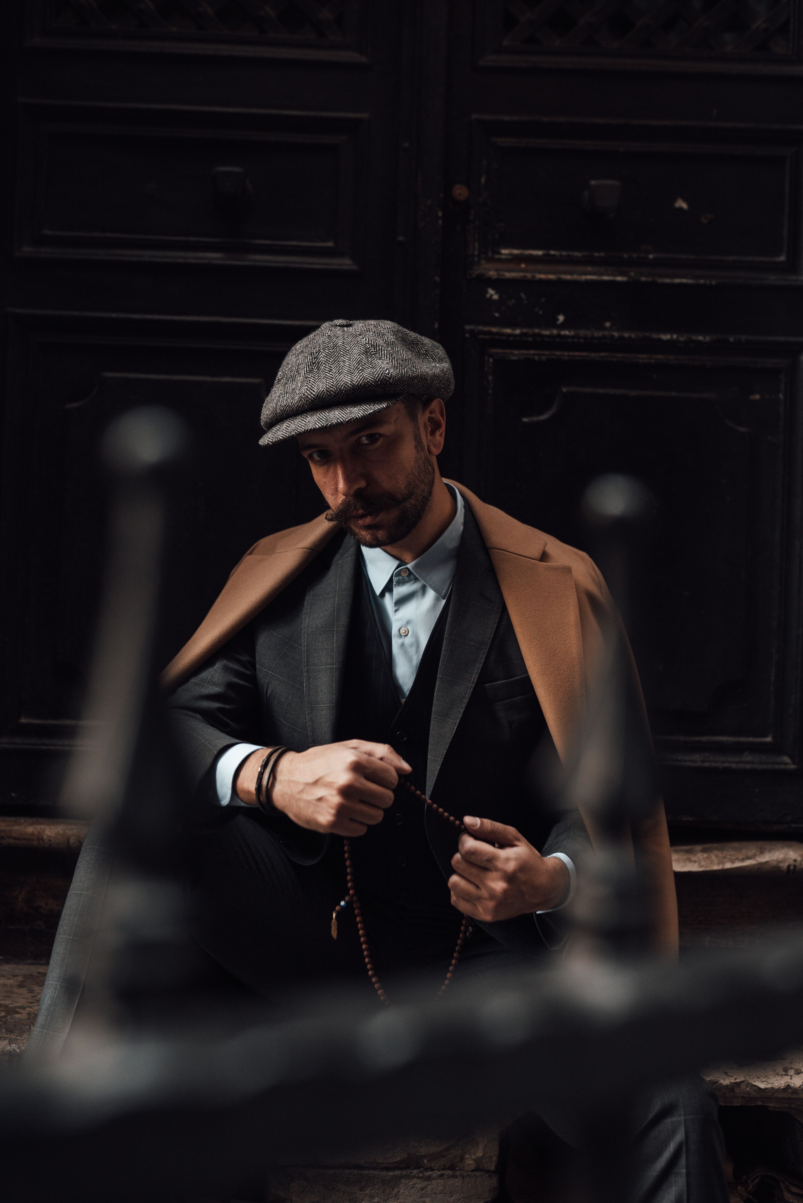 Serious male with beard and mustache in formal suit and stylish coat sitting on stairs of building and looking at camera in daytime