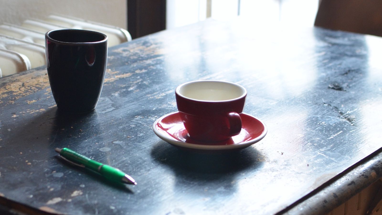 Two mugs and a green pen on a wooden table by a window