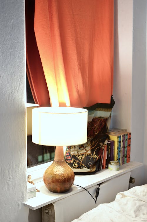 Glowing lamp near row of textbooks and ornamental curtain reflecting in window at home
