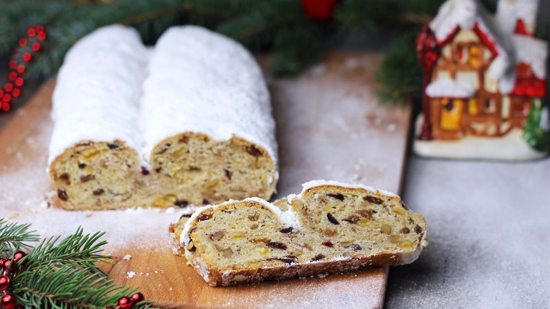 Zwei angeschnittene Christstollen mit dicker Puderzuckerschicht auf einem Holzbrett, davor eine Scheibe mit Rosinen, im Hintergrund Tannenzweige und ein Keramikhäuschen