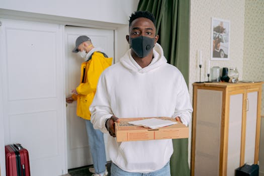 A masked delivery man holds a pizza box indoors. Another person enters through a door.