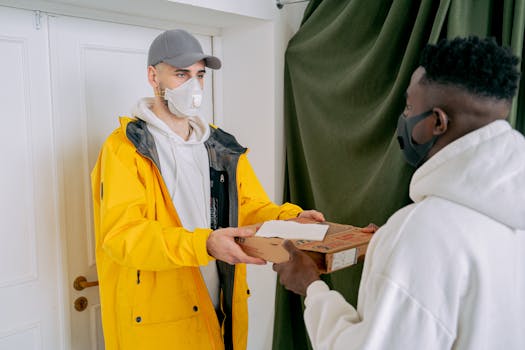 A deliveryman and customer exchange a pizza box wearing masks inside a home.