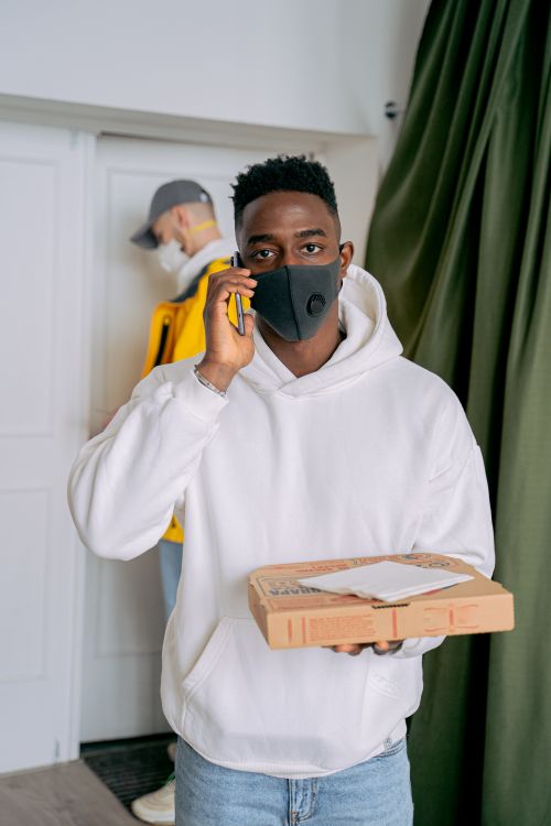 Free Man wearing a mask holds a pizza box and talks on the phone indoors. Stock Photo