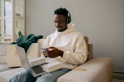 Free Man in a hoodie using a new smartphone and laptop on a sofa, indoors. Stock Photo