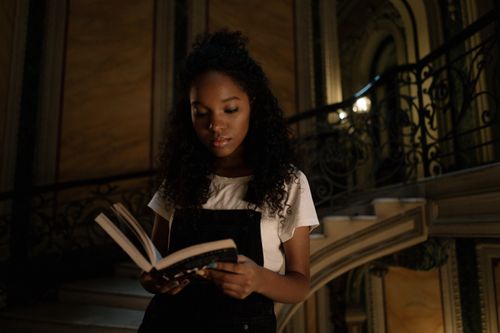 Free A young woman with curly hair reads a book while standing on an ornate staircase, immersed in thought. Stock Photo