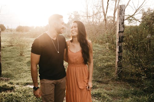 Content young ethnic couple looking at each other while standing close on lawn in countryside in back lit