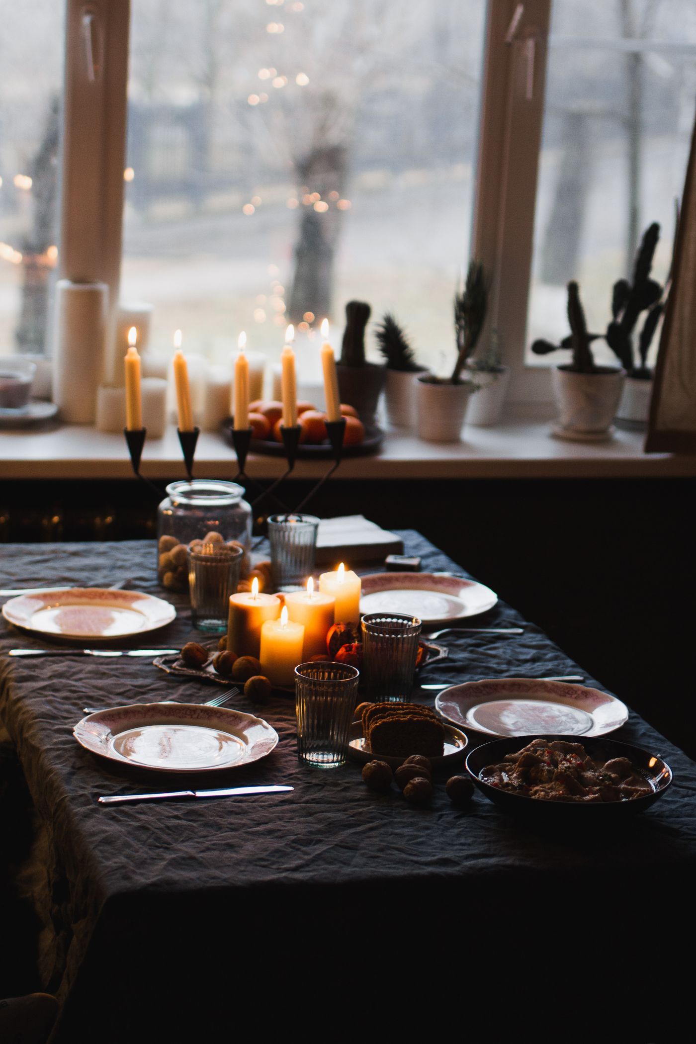 A warm, candlelit dinner table set beside a winter window