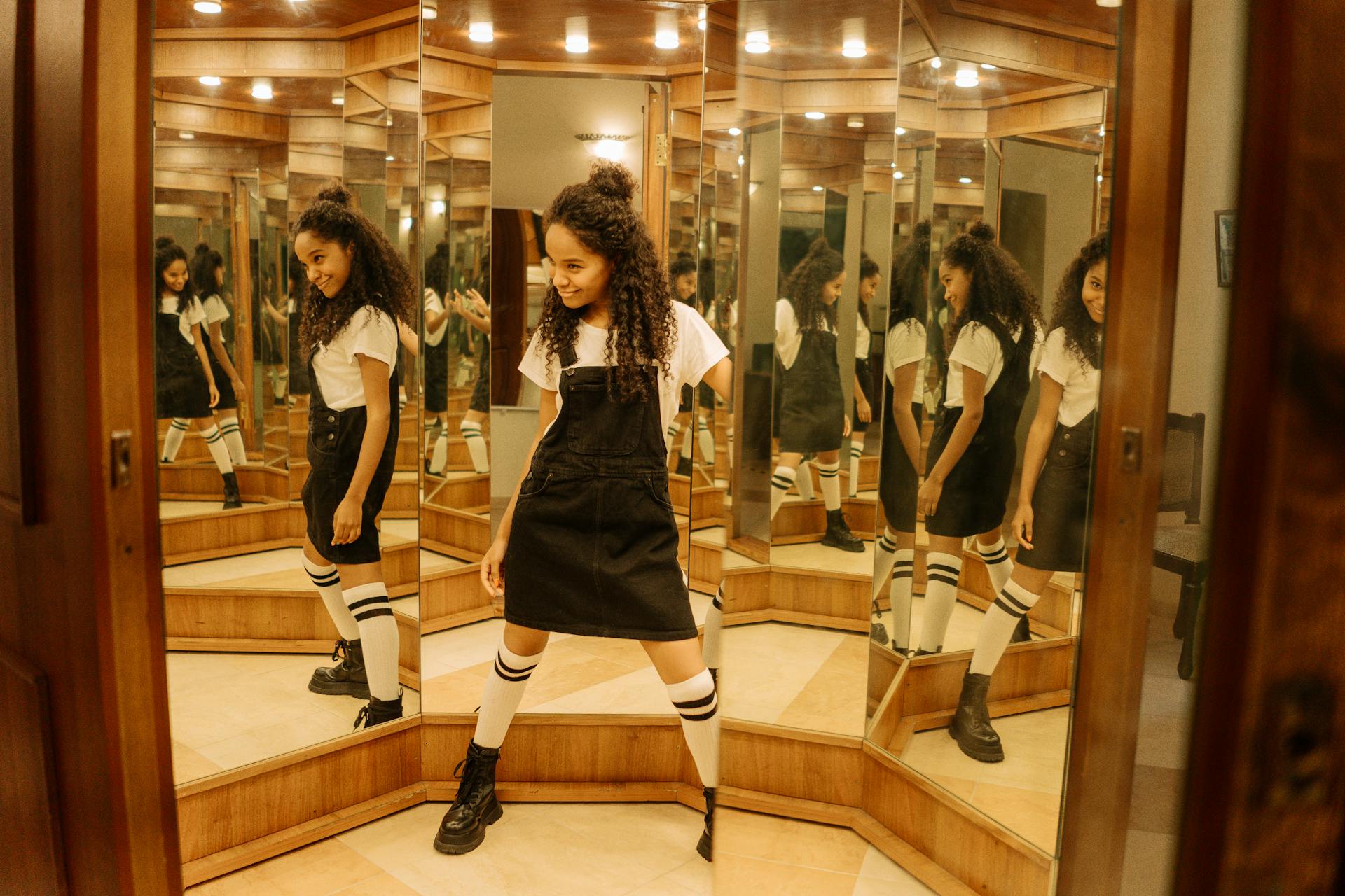 Teen girl with curly hair joyfully posing in a room with multiple mirrors.