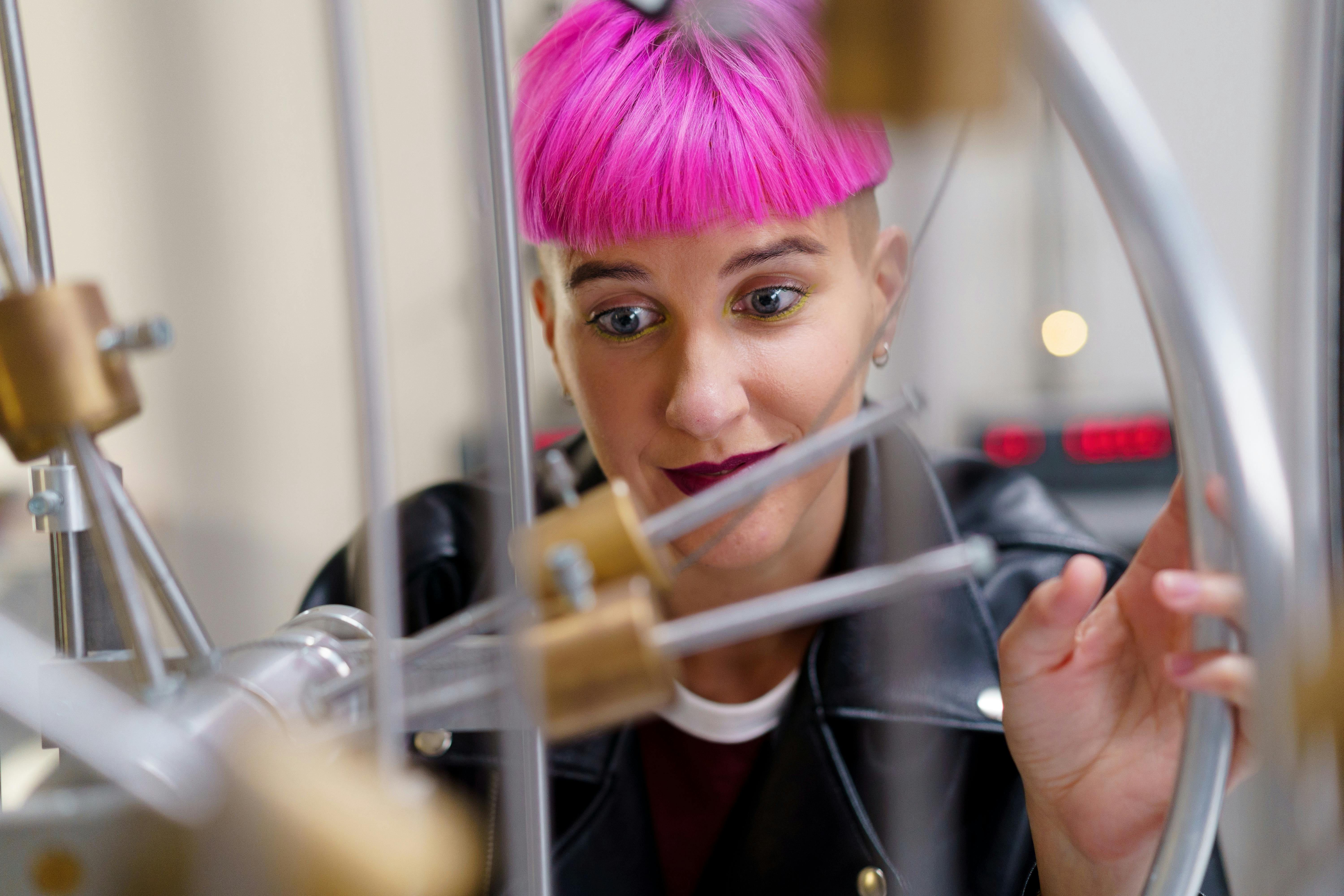 A curious young woman with pink hair examines scientific equipment in a lab setting.