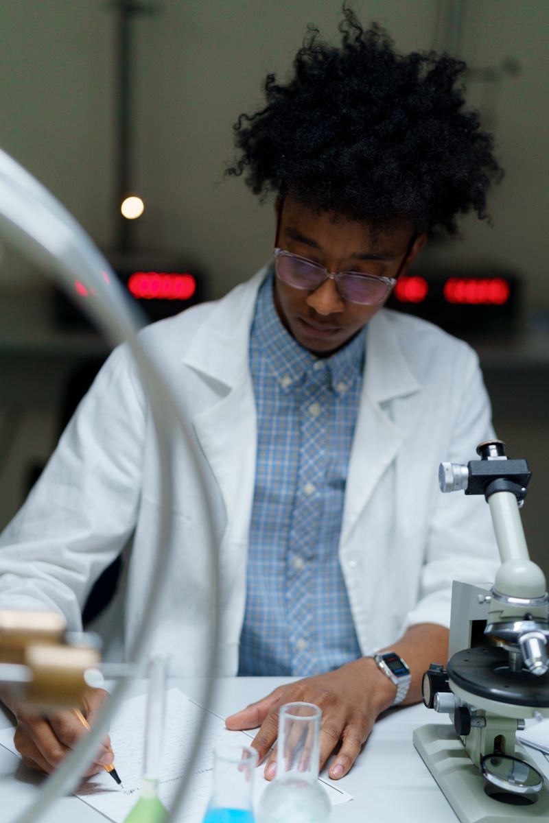 A Black lab scientist in a white coat and glasses works at a laboratory bench, writing notes beside a microscope, test tubes and glass flasks under low light.