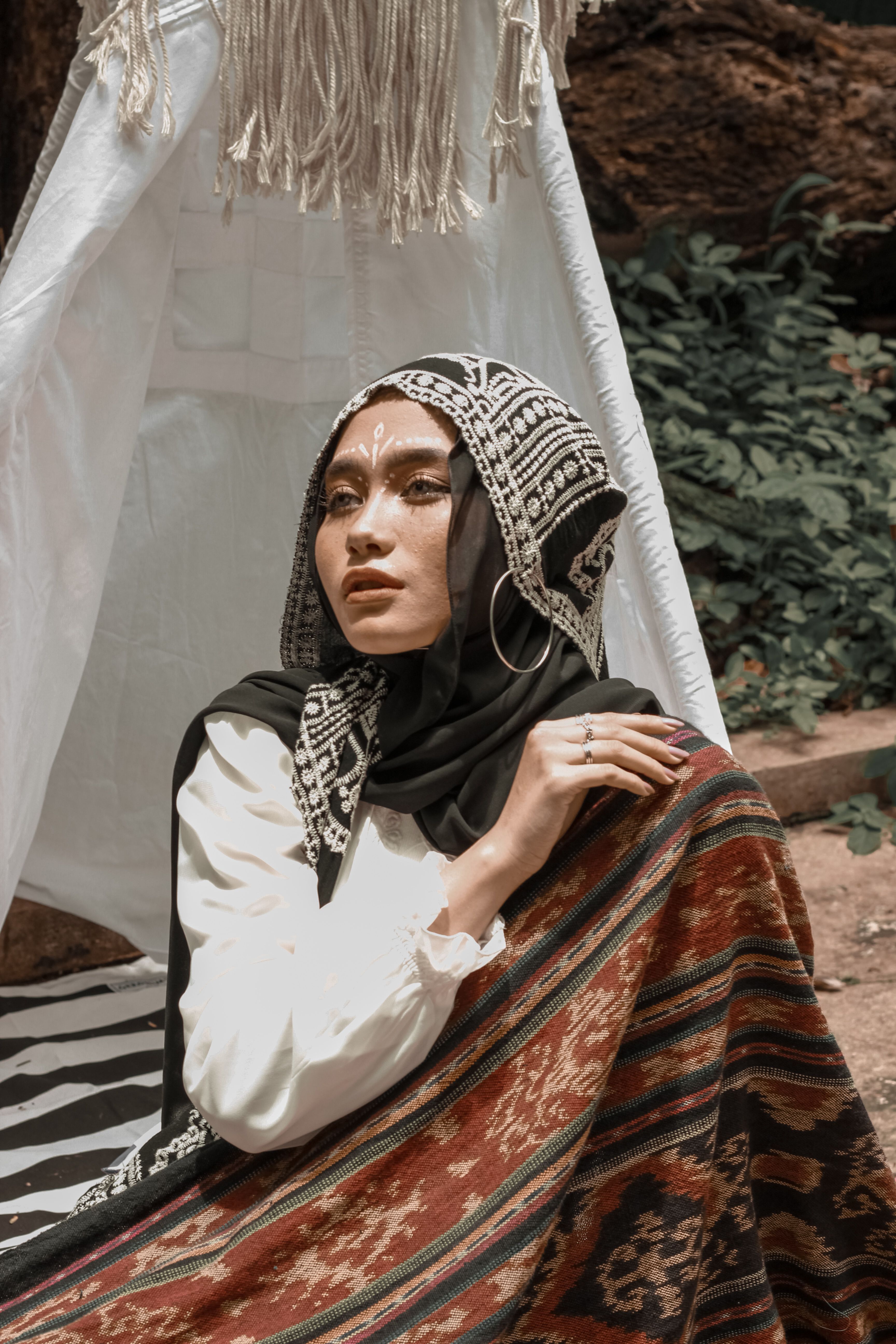 A woman in traditional attire with face paint sitting outdoors in a textile setting.