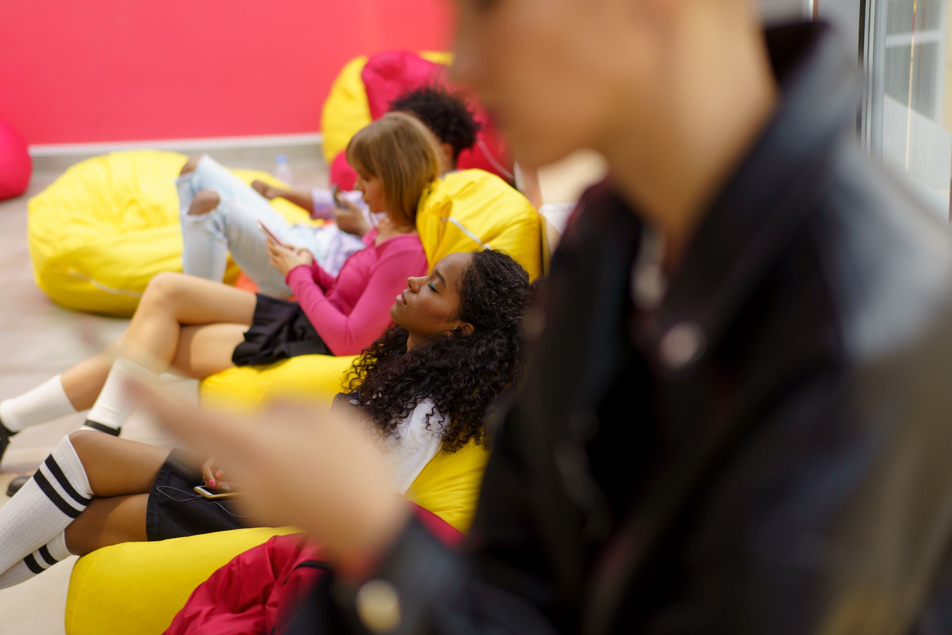 A group of teenagers relaxing on bright bean bags indoors, using smartphones and resting.
