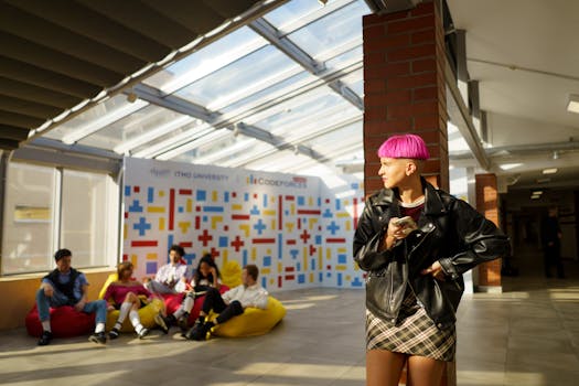 A young woman with pink hair and leather jacket stands confidently in a sunlit university lounge with peers in the background.