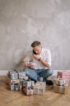 Man sitting on wooden floor talking on phone, surrounded by colorful gift boxes against gray wall.