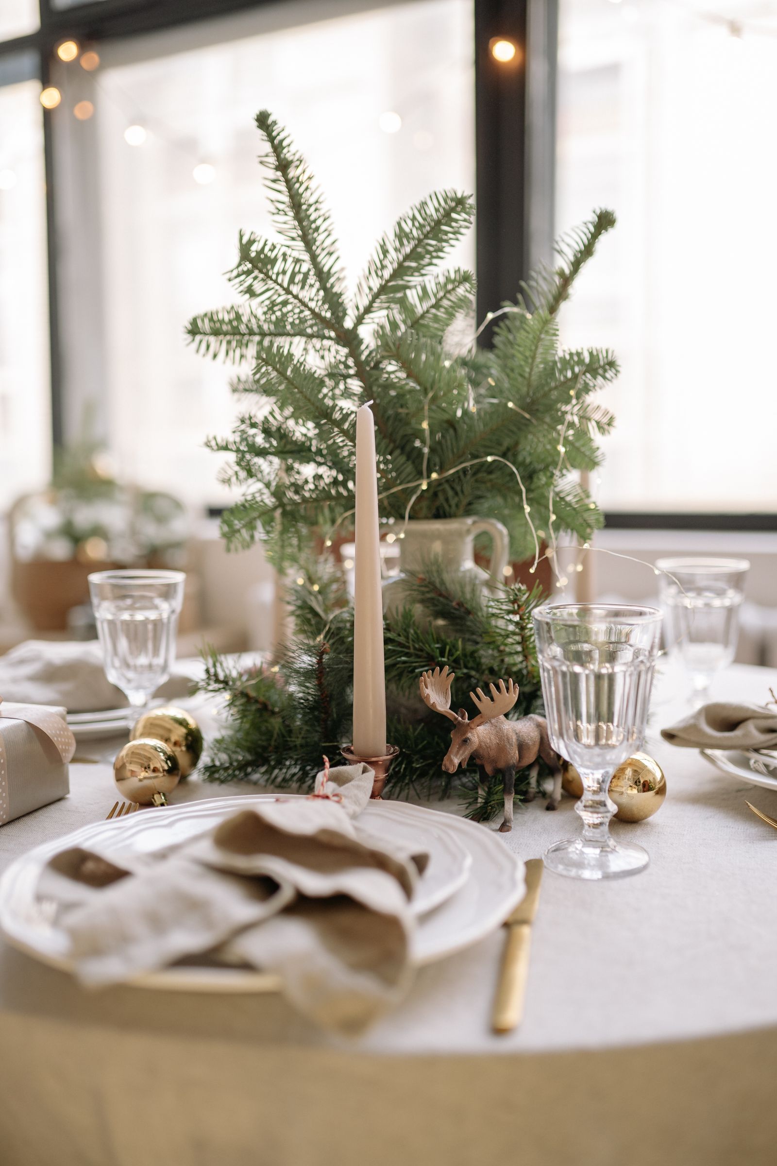 Festive dining table with a white tablecloth, ordinary place settings, greenery, and restrained Christmas decorations.