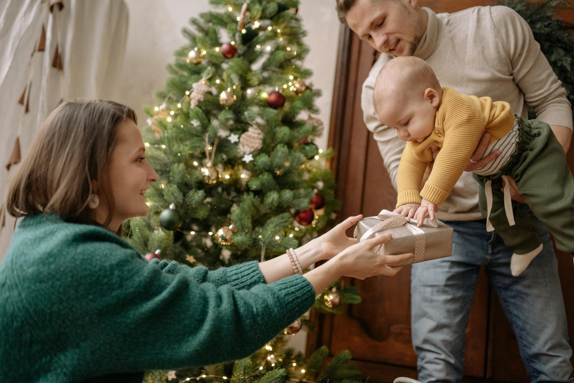 A joyful family moment with parents exchanging a gift with their baby by the Christmas tree.