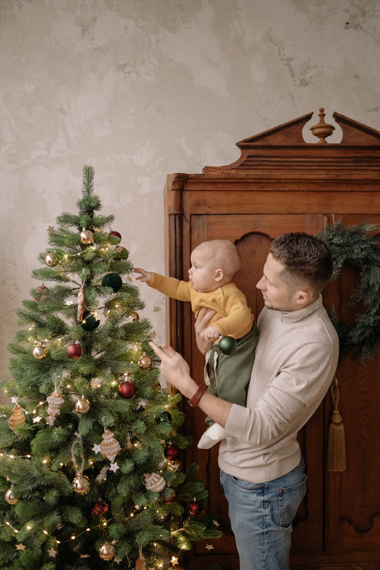 Man Holding A Baby Touching A Christmas Tree And Wooden Wardrobe Behind