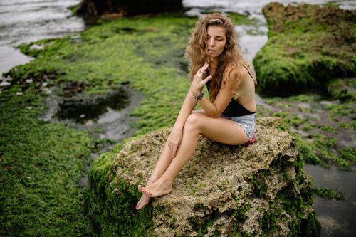 Free Woman with curly hair sitting on a moss-covered rock by the seaside, relaxing outdoors. Stock Photo