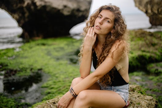 A woman in casual summer attire sitting by a rocky seashore, exuding a relaxed vibe.