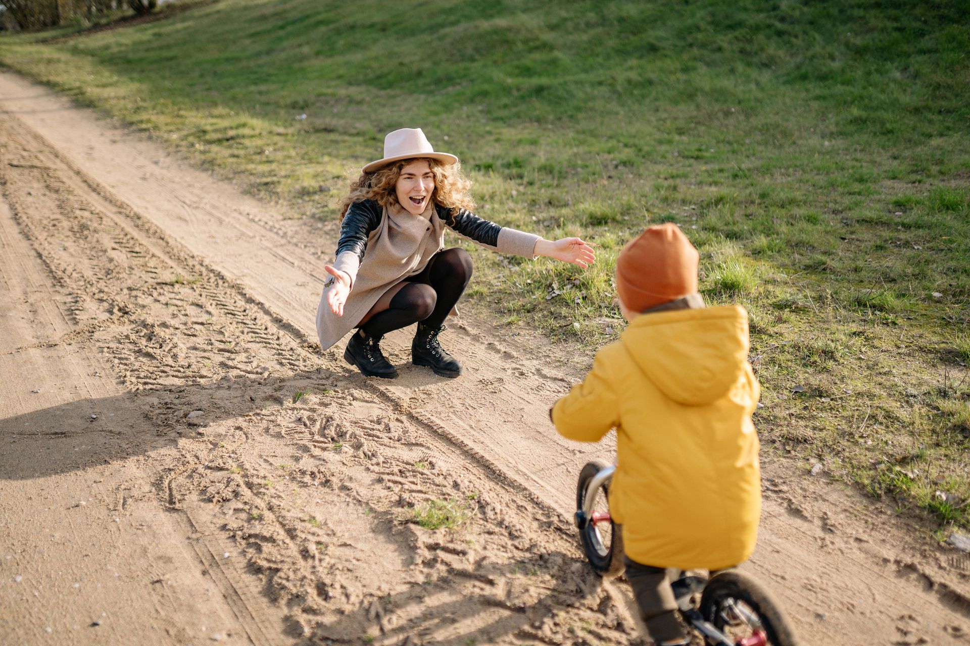A mother crouching down with arms open as her young son rides towards her on a balance bike