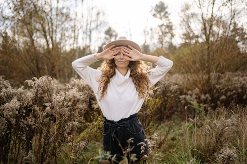Free Woman in a hat playfully covering her eyes in an autumn field, showcasing nature and fashion. Stock Photo