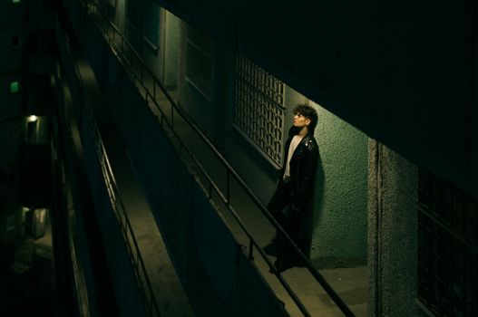 A man in a leather jacket stands on a moody, dimly lit balcony at night, evoking solitude.
