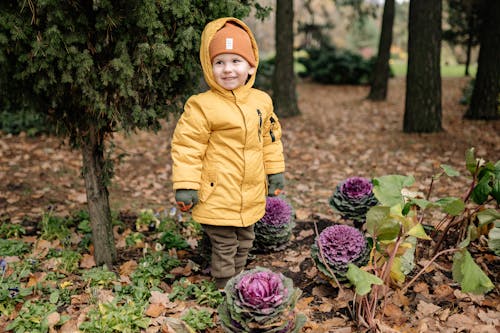Child in Yellow Hoodie Jacket and Yellow Knit Cap Standing Near Plants With Flowers