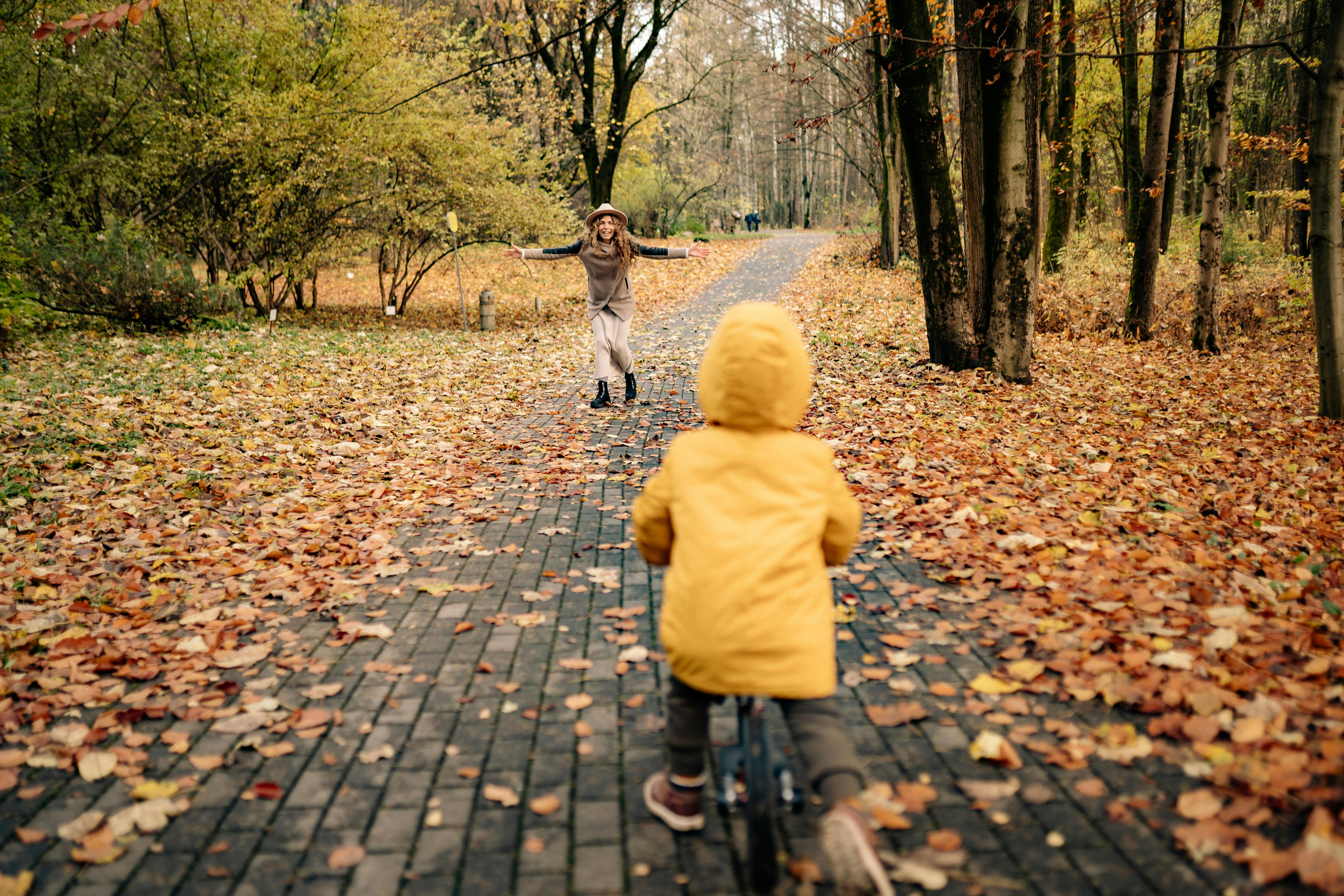 A child in a yellow jacket rides a bicycle towards a mother in a park during fall season.