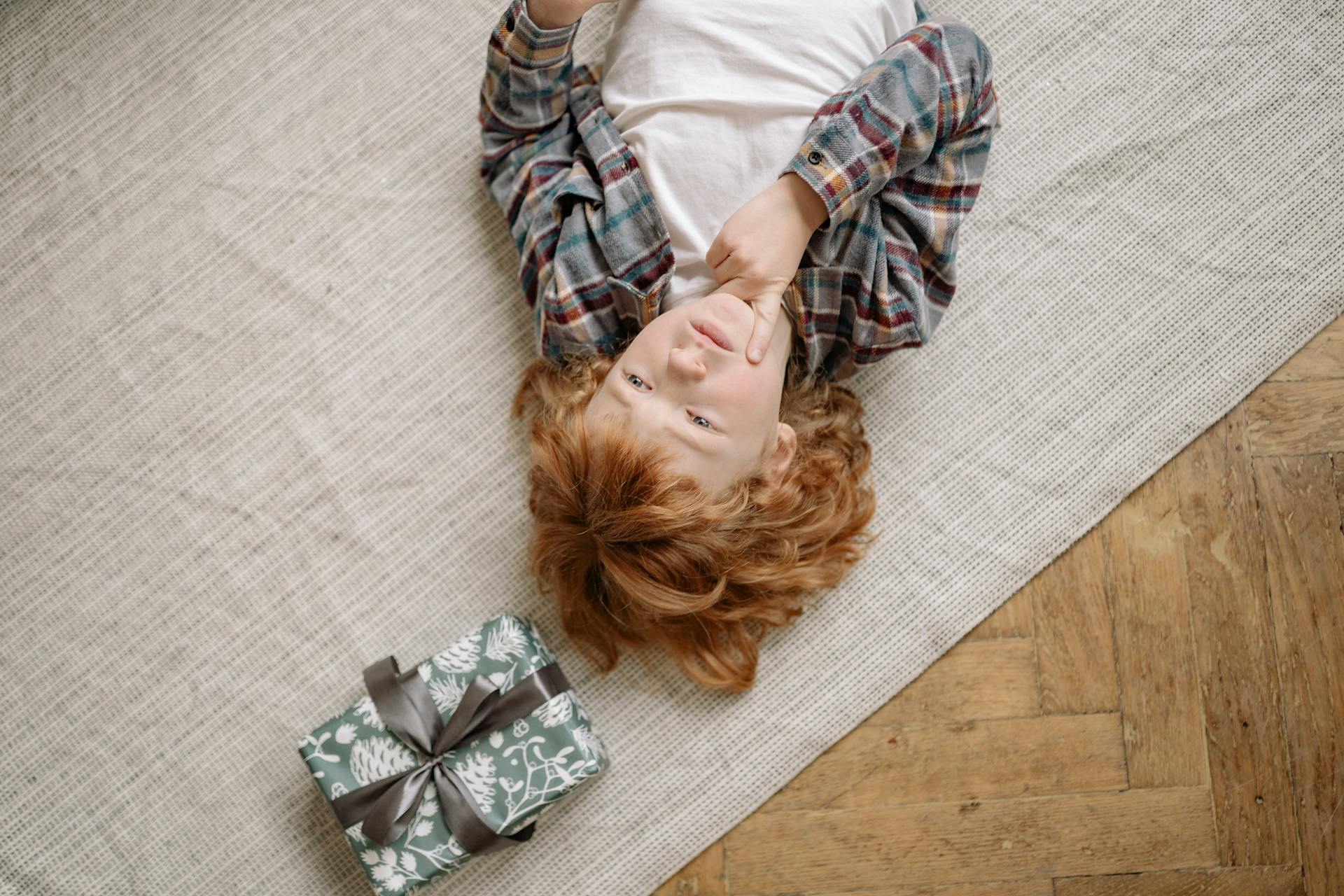 Thoughtful child lying on the floor next to a wrapped gift indoors.