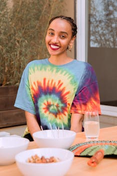 A cheerful woman in a vibrant tie-dye shirt sits indoors, smiling at a dining table.