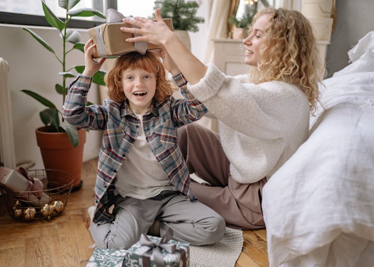 A Kid Sitting On The Floor With A Woman Holding A Box With Gray Ribbon On His Head