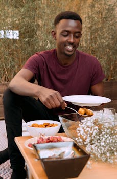 A young man happily serves himself food at an outdoor table setting.