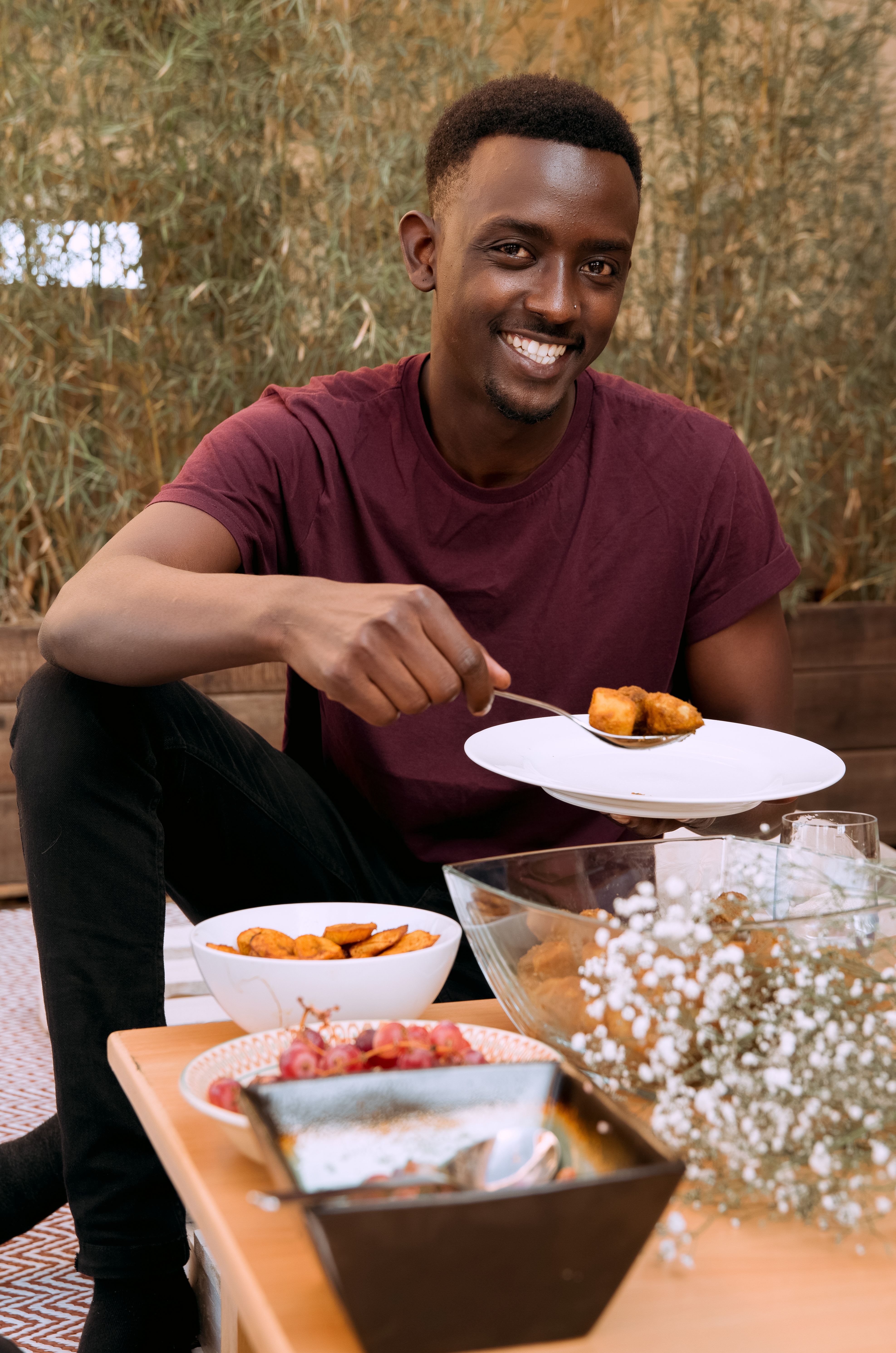 A smiling man holds a plate of food while sitting at a table indoors.