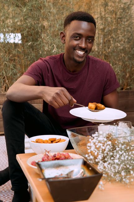 A smiling man holds a plate of food while sitting at a table indoors.