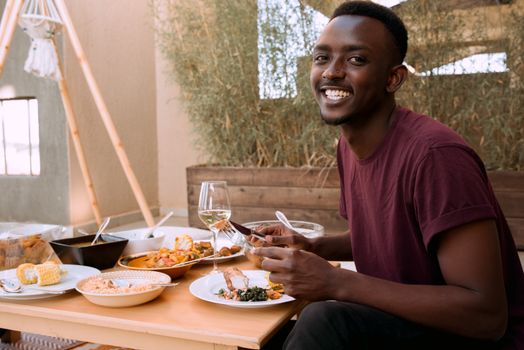 A young man happily enjoys a meal outdoors at a table filled with various dishes and a glass of white wine.