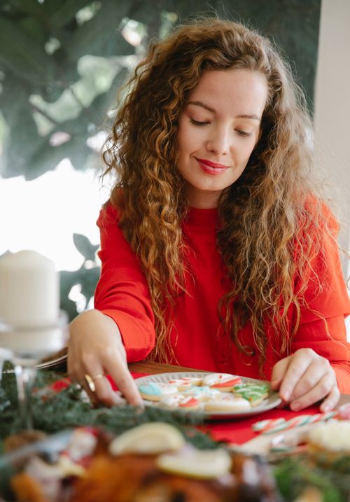 Free Positive female sitting at table with various food and with plate full of cookies with icing during Christmas  holiday celebration Stock Photo