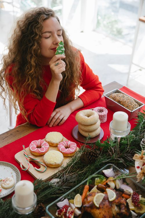 Free From above of pleased female with closed eyes smelling fresh cookie while sitting at table with doughnuts meat and coniferous branch Stock Photo