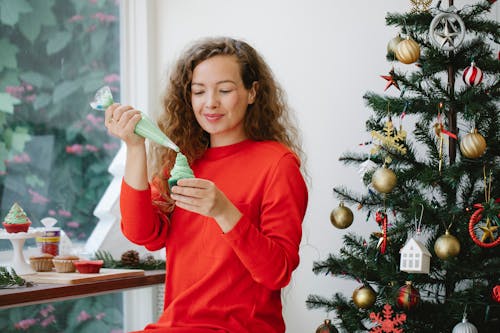 Cheerful female with pastry bag piping green cream on cupcake while sitting near window and decorated fir during Christmas holiday preparation