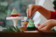 Unrecognizable female sprinkling topping on green frosting of cupcake while standing near windowsill with wooden board and coniferous branch on blurred background